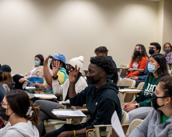 A group of students sit at desks in a classroom, with several students raising their hands to answer a professor's question