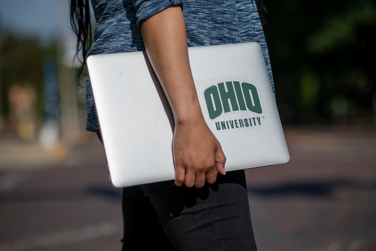 Student's arm for the elbow down holding a laptop with an Ohio University sticker