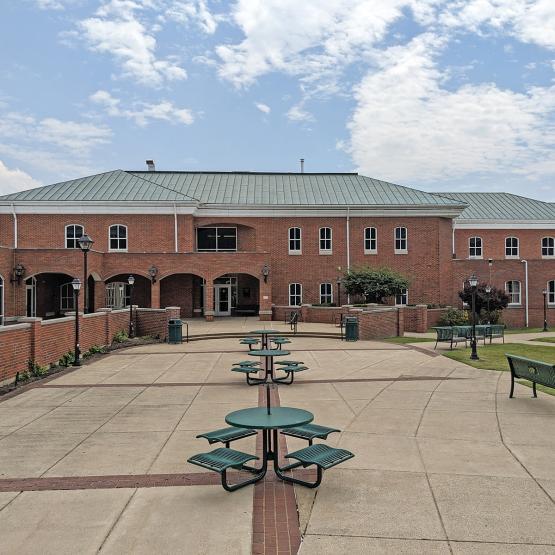 A wide two-story brick building with a pale green tiled roof. There is a courtyard with green tables to sit and eat at.