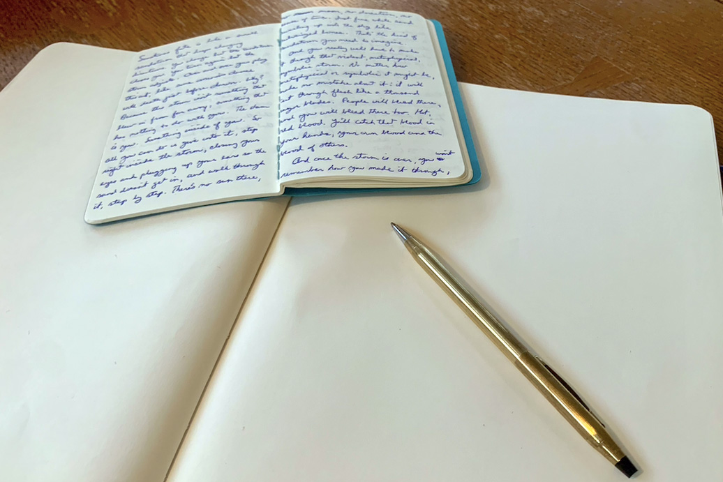 A notebook with writing lays on top of a blank notebook by a pen.