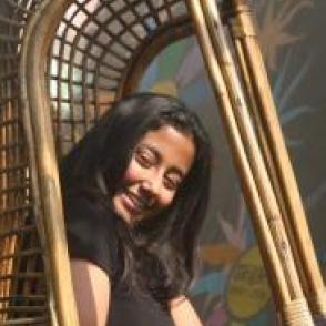 Aanya Datta sits in a wicker chair, smiling at the the camera in a black t shirt.