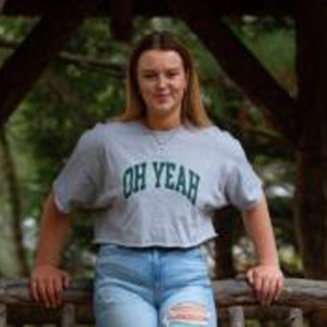 Annika Lindsey leaning back on a post, wearing a gray shirt that says 'oh yeah' in green lettering. 