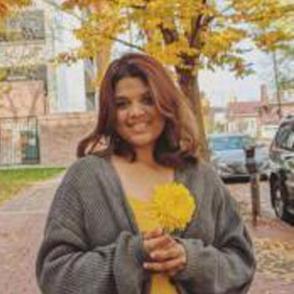Ankita smiles at the camera in front of a yellow-leaved tree, in a yellow shirt of a similar color with a grey cardigan over it.