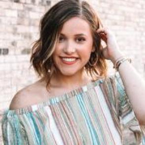 Delaney Brander tucks hair behind her ear, smiling at the camera. She wears a vertically striped shirt that is teal, tan, and white.