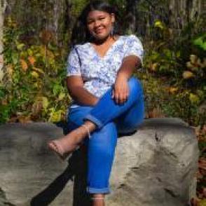 Giah sits with one leg crossed over the other, wearing a white a blue floral shirt and deep blue jeans.