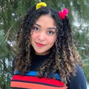 Judinya Thwaites-Bank stands, putting her hands in her back pockets. She wears a dark blue shirt that has four thick stripes; the stripes are blue, red, orange and yellow. Her hair is in pigtails, the left scrunchie is yellow, while the right scrunchie is red.