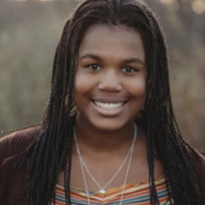 Maellie-Jade Jean-Francois smiles at the camera, wearing a horizontally striped shirt that is yellow, orange, cream and blue. Over her shirt she wears a deep brown cardigan.