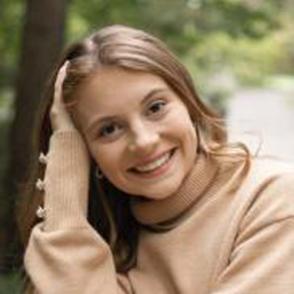 Riley Sargent leans on one arm, smiling at the camera and wearing a cream sweater with buttons on the cuff.