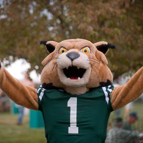 Bobcat mascot wearing a green sports jersey with the number 1 on the front, standing outdoors with arms raised in a celebratory pose.