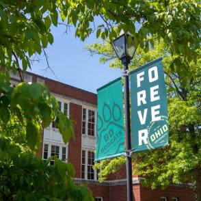 Two green banners on a lamppost reading ‘Go Bobcats’ with paw prints and ‘Forever Ohio,’ displayed near a brick campus building surrounded by trees.