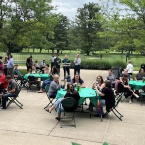 Outdoor event with people seated at green-covered tables and others standing on a patio surrounded by trees.