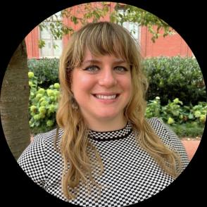 a woman smiling with long blondish brown hair and bangs wearing a black and white patterned top in an outdoor setting