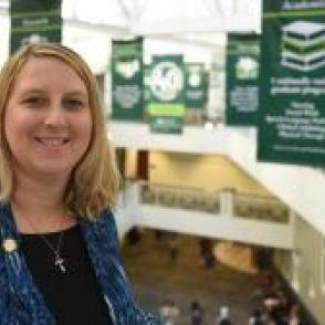 a woman with shoulder length blond hair standing in front of an atrium balcony wearing a black shirt and jeans jacket smiling. 