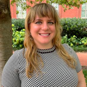 A woman smiling with long blondish brown hair and bangs wearing a black and white patterned top in an outdoor setting