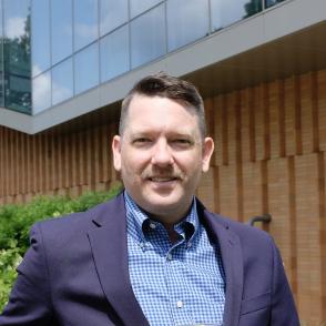 a headshot of a white male with short brown hair smiling facing the screen. wearing a dark blue suit jacket and a light blue shirt 