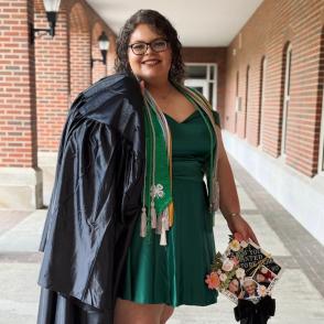 Woman standing in graduation gown holding cap and smiling