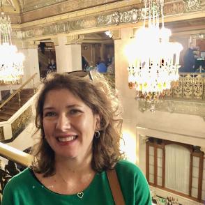 image of a white woman with light colored hair in a green top in a gold detailed room with a crystal chandelier behind her