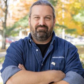 image of a white male with dark hair with grey and a beard with his arms crossed in front of him wearing a blue denim shirt with cuffs rolled. 