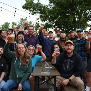 Group of people sitting and standing around a table holding drinks up in cheers.