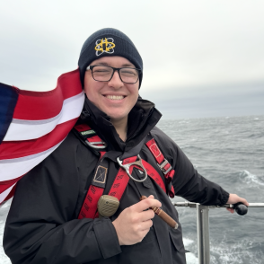 image of a make standing in font of a red and white striped flag wearing a black hat and a black jacket on the deck of a boat with the ocean behind it