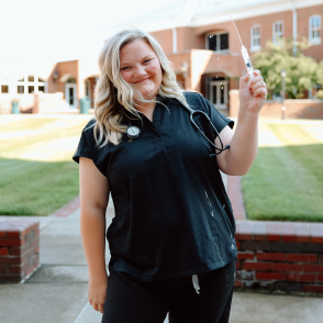 Graduate in scrubs smiling in front of OHIO Southern wearing stethoscope and squirting syringe 