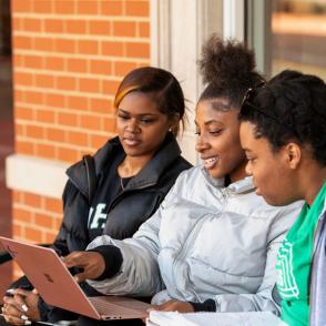 peer educator sits with two students talking smiling and pointing to a laptop screen