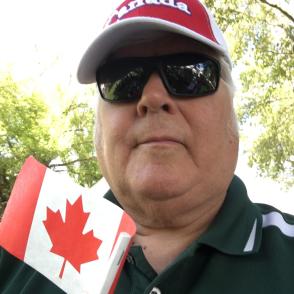 a man wearing a red Canada hat, sunglasses, and a dark polo holds a small Canadian flag in front of him