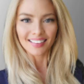 a blond woman with her hair down faces the screen smiling wearing a dark blue top 