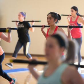 group of students hold bars over their shoulders as they do crisscross jumps in pilates class 