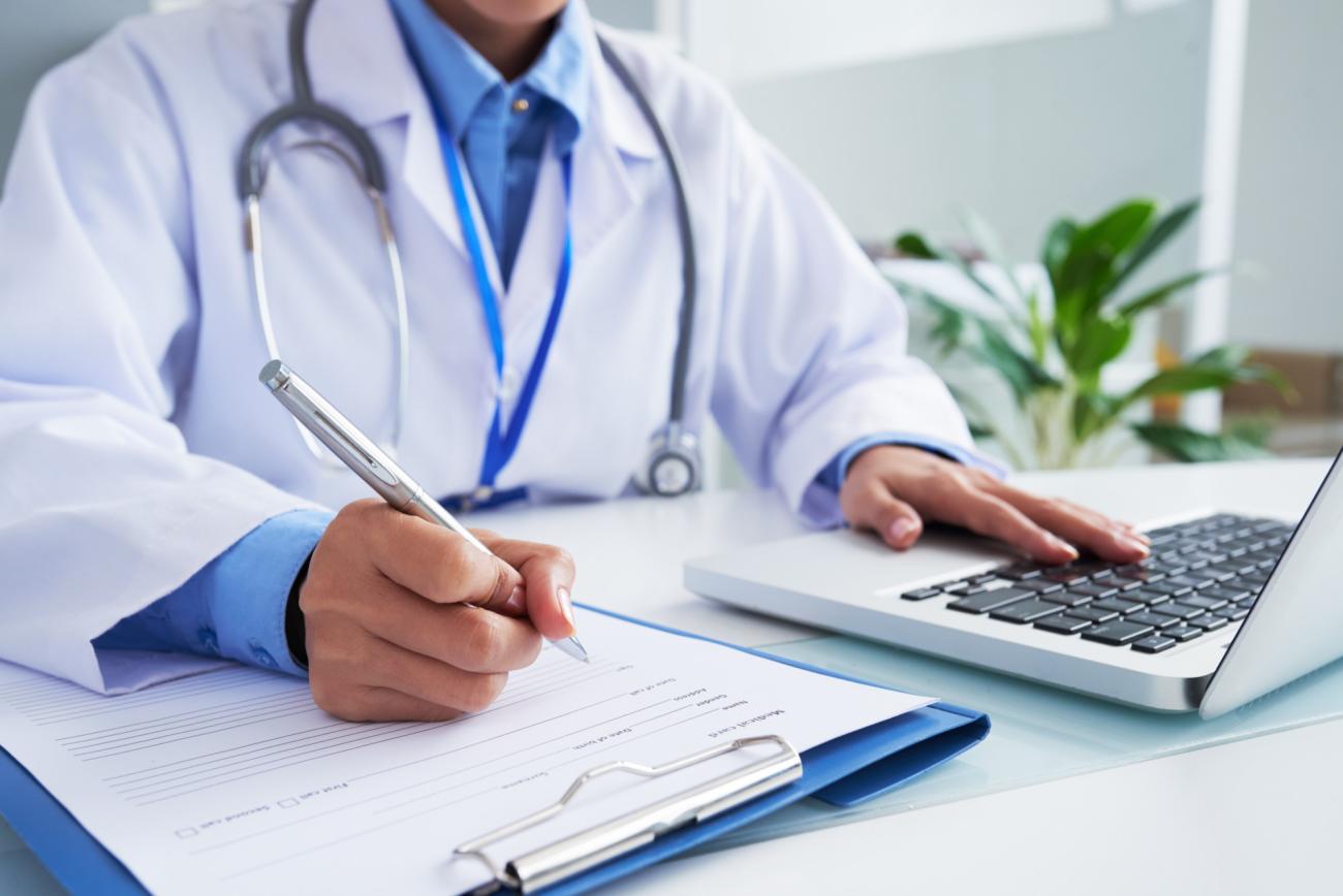 Doctor writing on form and typing on laptop keyboard. Image by Freepik.