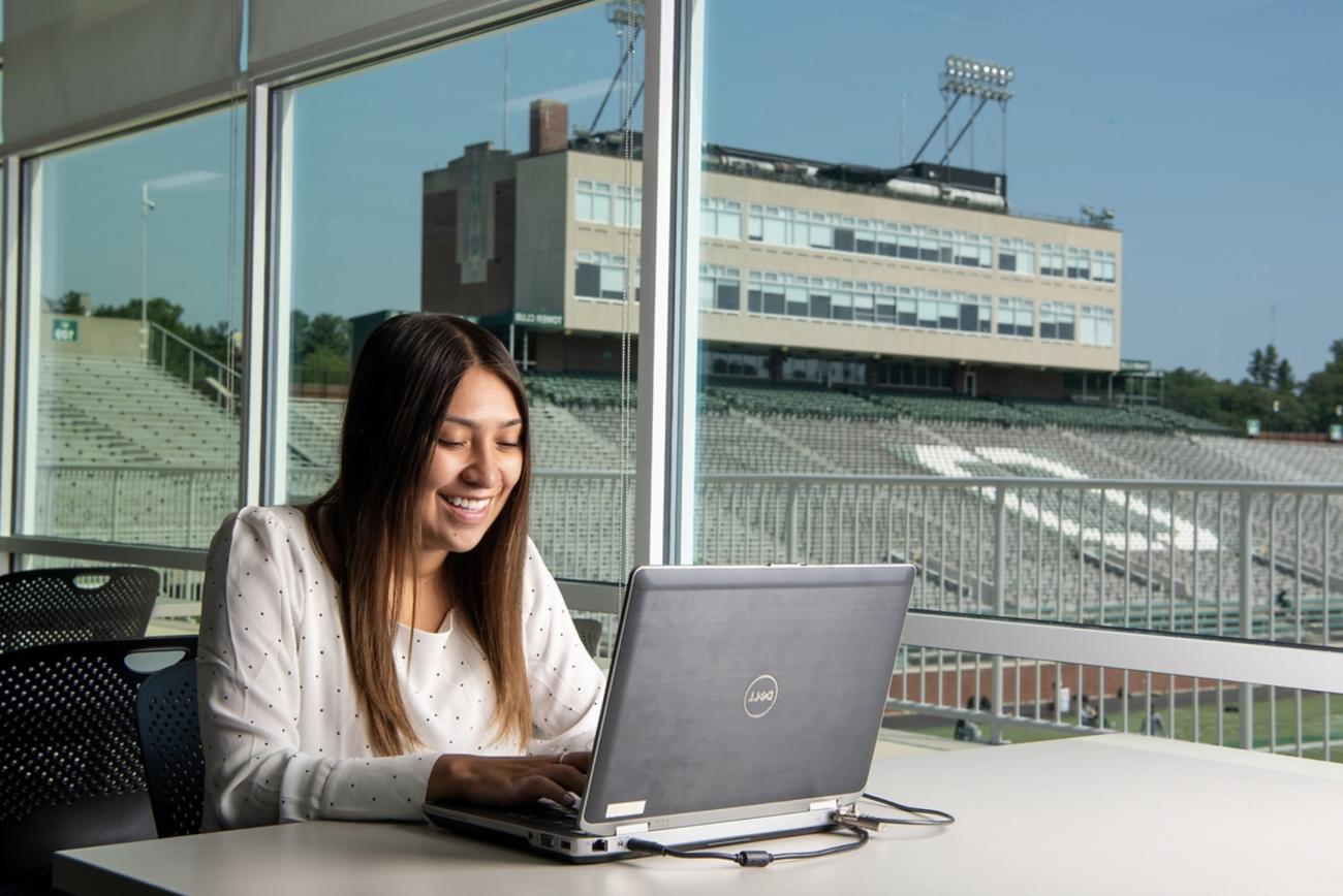 A woman smiles and types on her computer, with Peden Stadium in the background. 