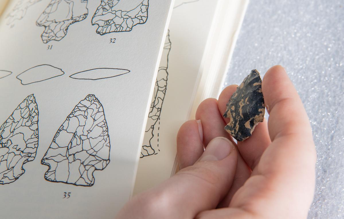 Closeup of a hand holding an excavated arrowhead next to a diagram