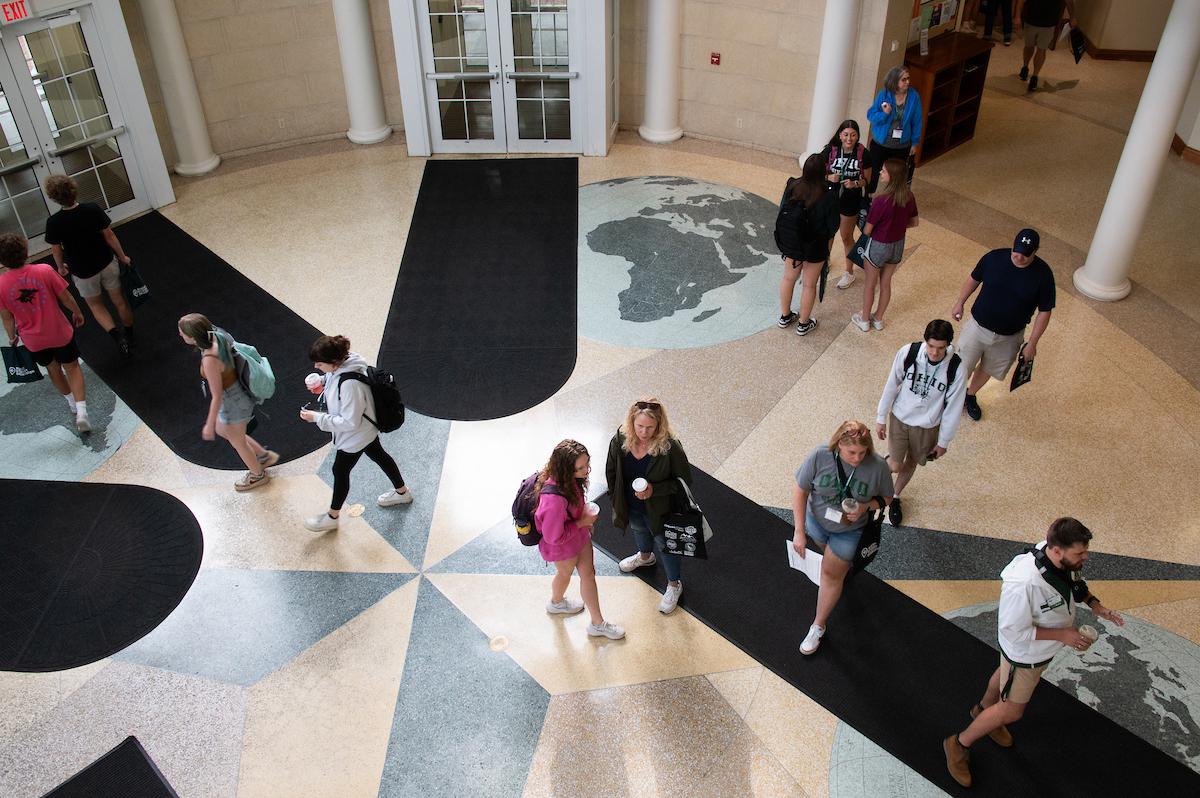 The photo is taken from above, students and their supporters walk through an atrium on the fourth floor of baker wearing Bobcat Student Orientation lanyards. They are surrounded by doors with black rugs leading to them. 