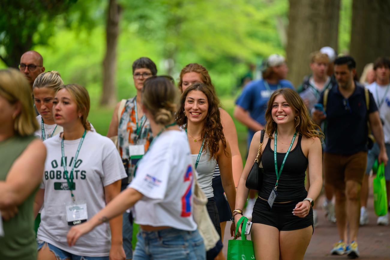 A group of students walks along a brick path surrounded by trees. The focus of the photo are two students facing forward smiling while listening to one student who is turned around, presumably speaking to them. 