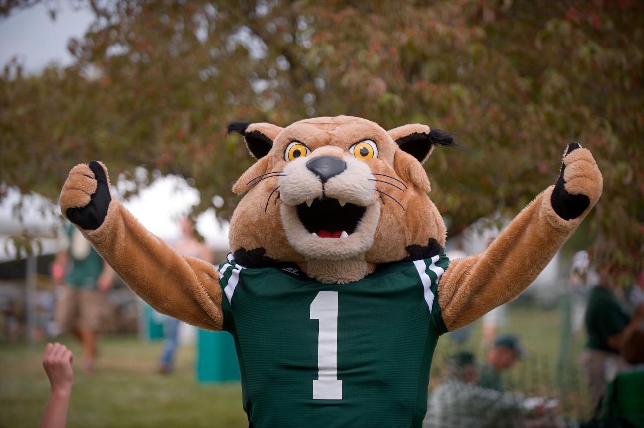 Bobcat mascot wearing a green football jersey with white stripes and the number 1, standing outdoors with arms raised in a celebratory pose. The background shows blurred trees and people, suggesting a sports or school event.