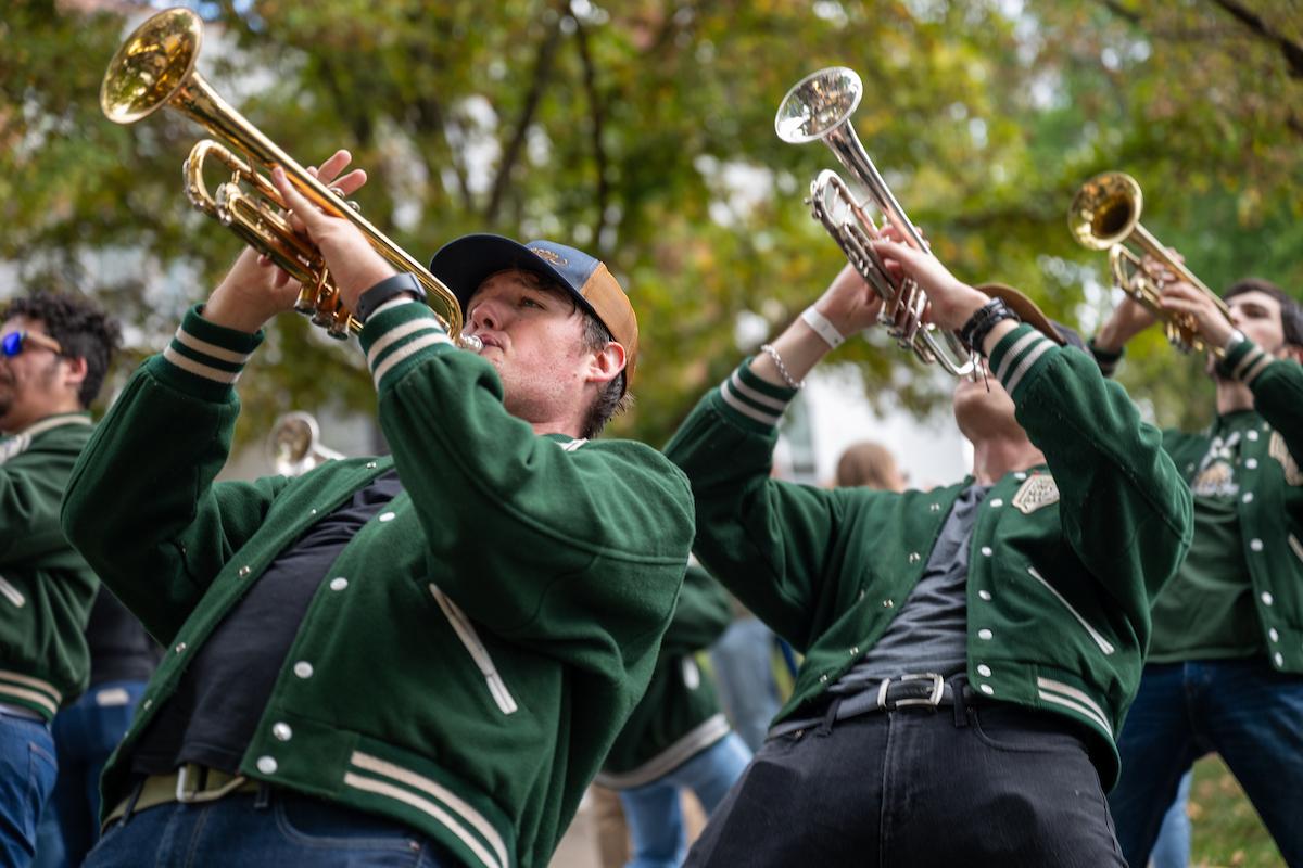 Marching band members wearing green jackets perform outdoors, playing brass instruments while leaning back in formation under trees.