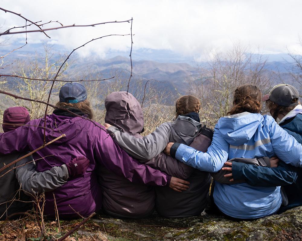 Students sit together with arms linked across their backs as they take in the view after a hike.