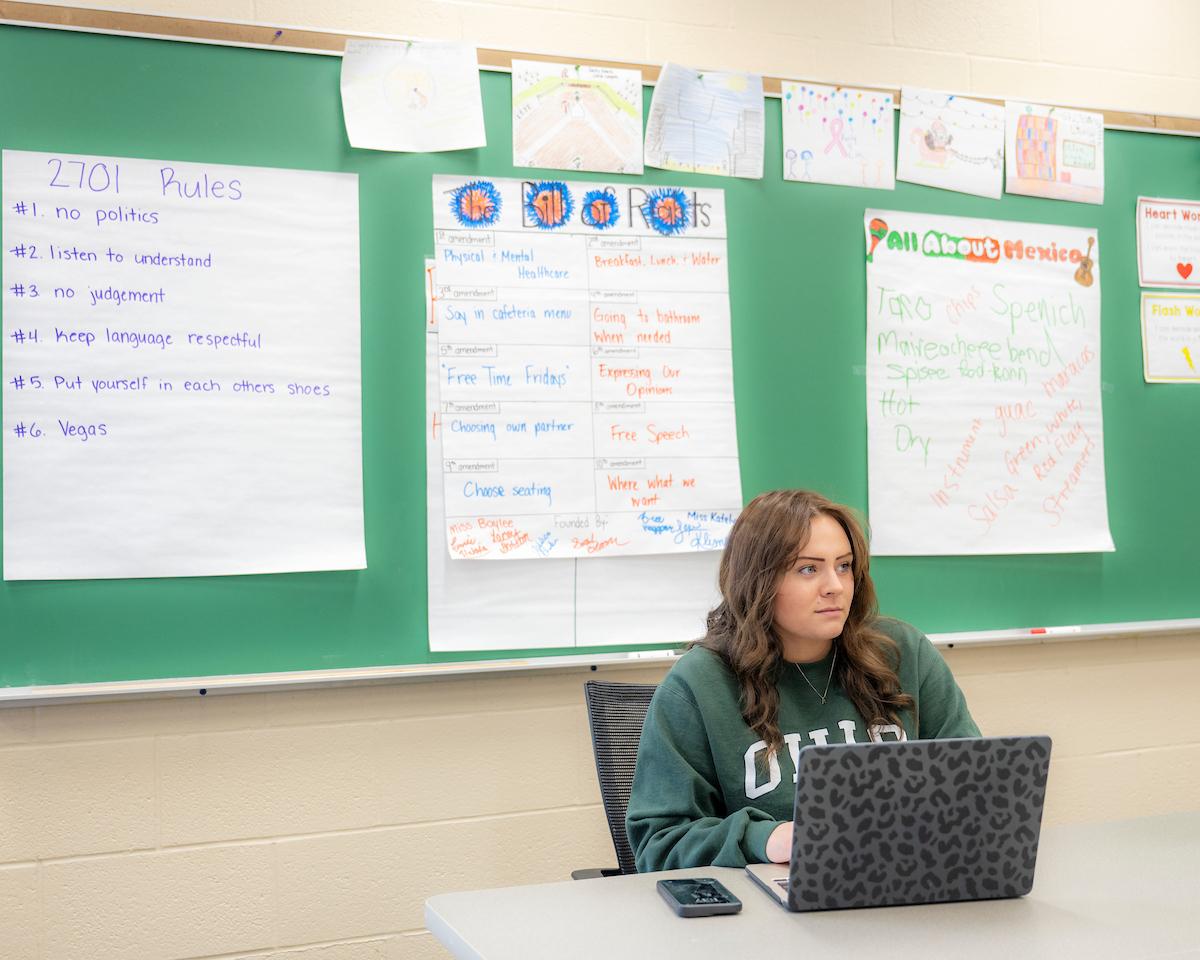 Teacher in a classroom sitting at a desk looking at laptop