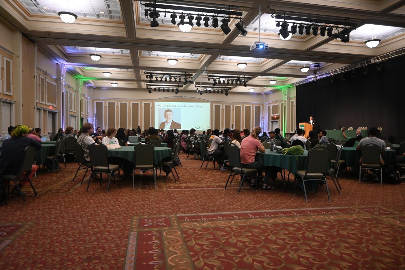 A wide shot of a room filled with people listening to a speaker