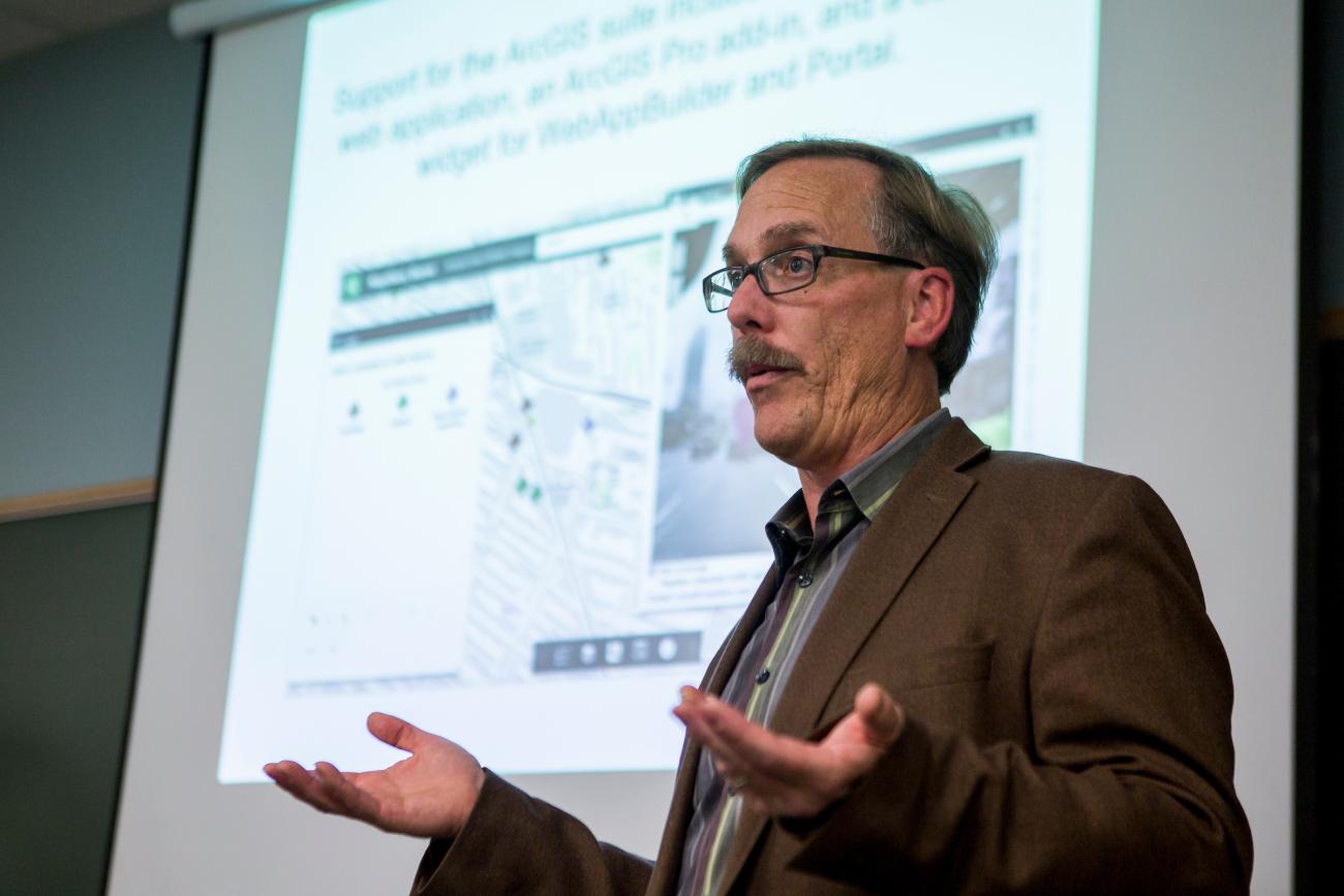 Man presenting to a classroom with a slideshow slide on projector screen behind him