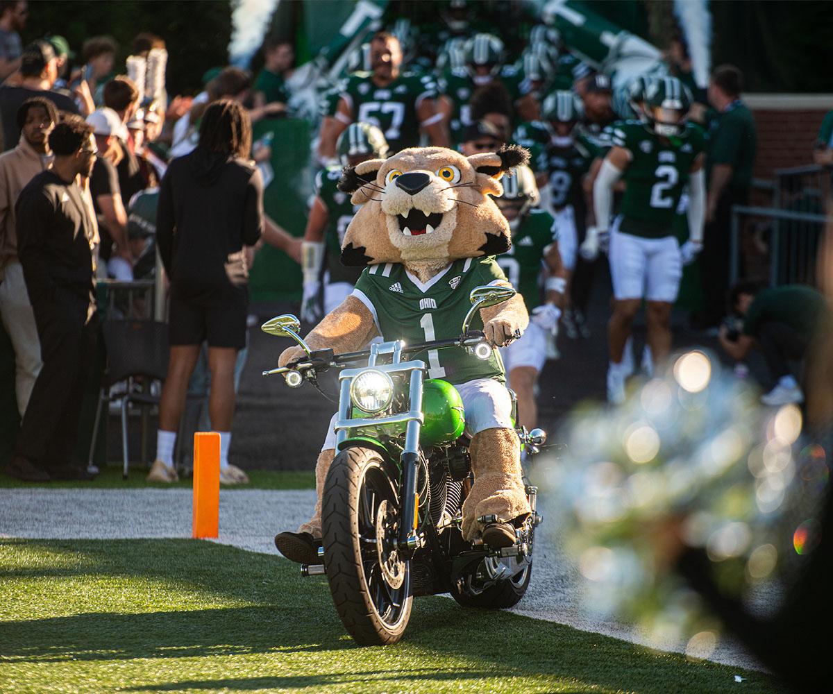 Rufus the Bobcat, OHIO's mascot, rides a motorcycle on the football field in front of football players.