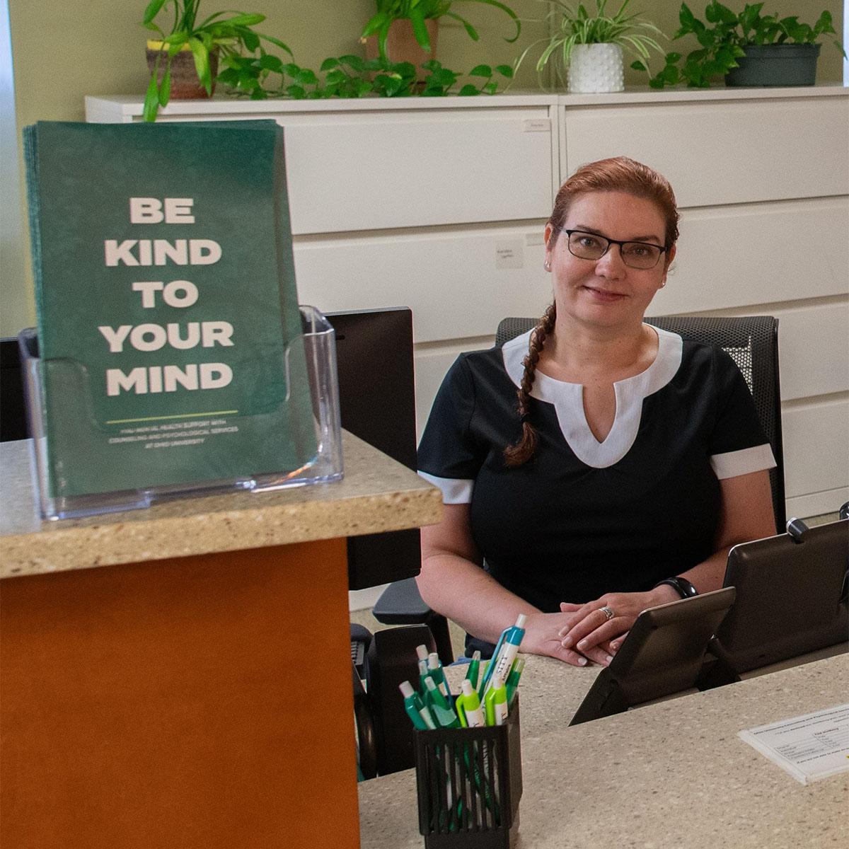 A front desk worker at CPS smiles next to a pamphlet that says, "Be kind to your mind."