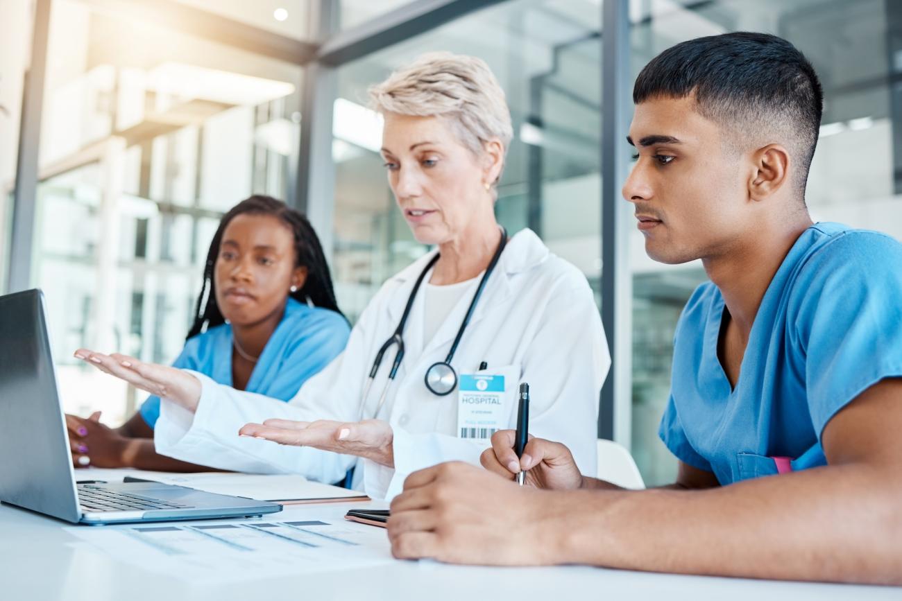 nurse in white coat teaching 2 nurses in blue scrubs