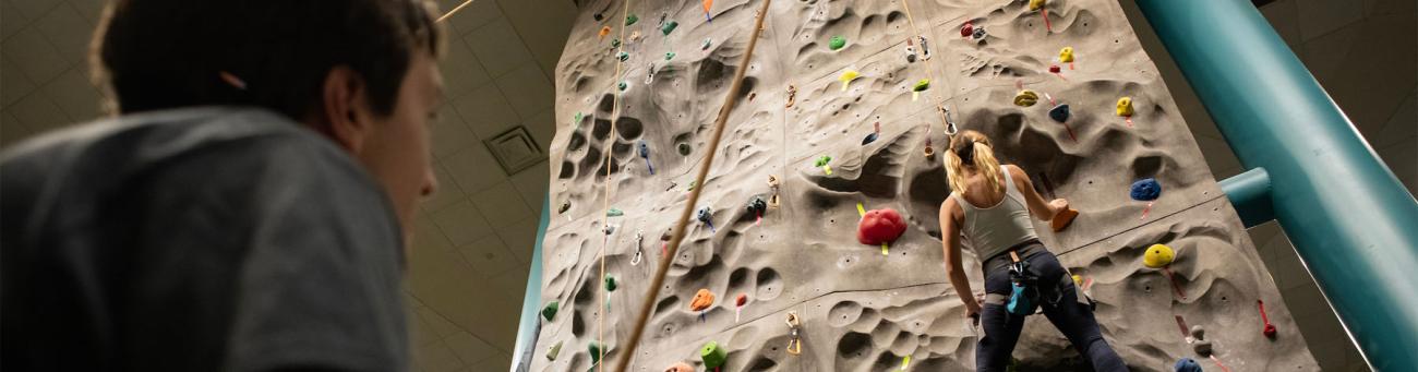 man looks at the climbing wall from below while a woman climbs it 