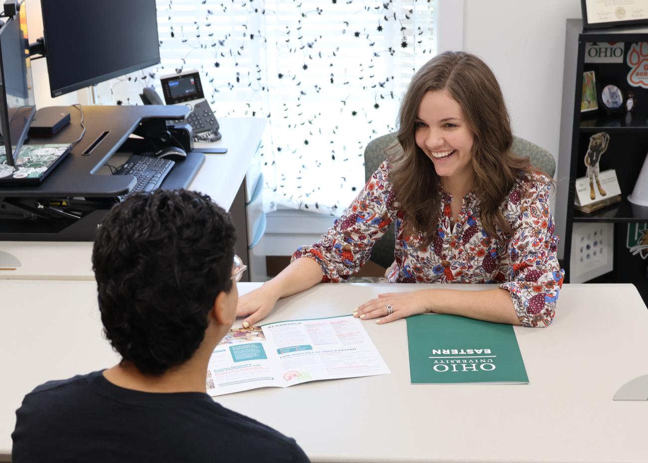 Woman smiling facing a student, both are sitting facing each other at a desk inside an office