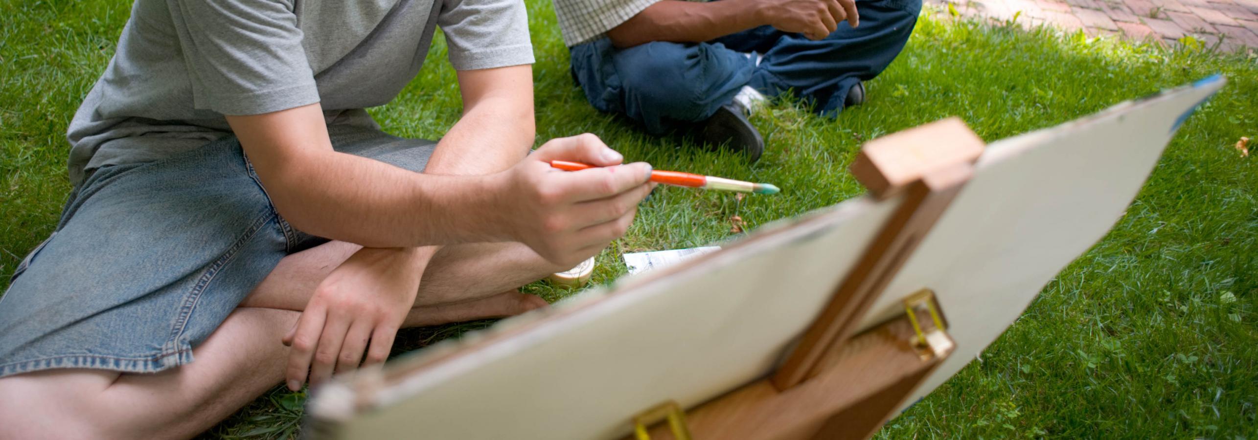 Two students sit outside painting on an easel.