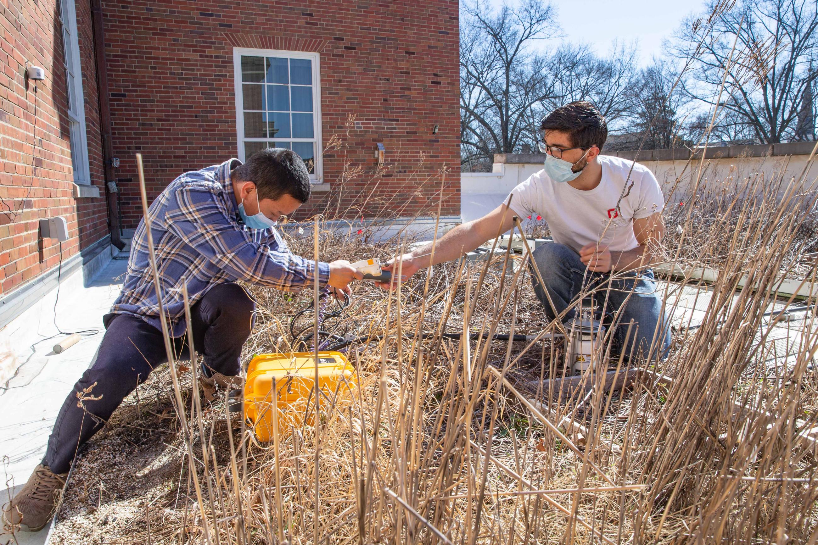 Researchers handle electronic instruments while kneeling in a plant bed on the Schoonover green roof.