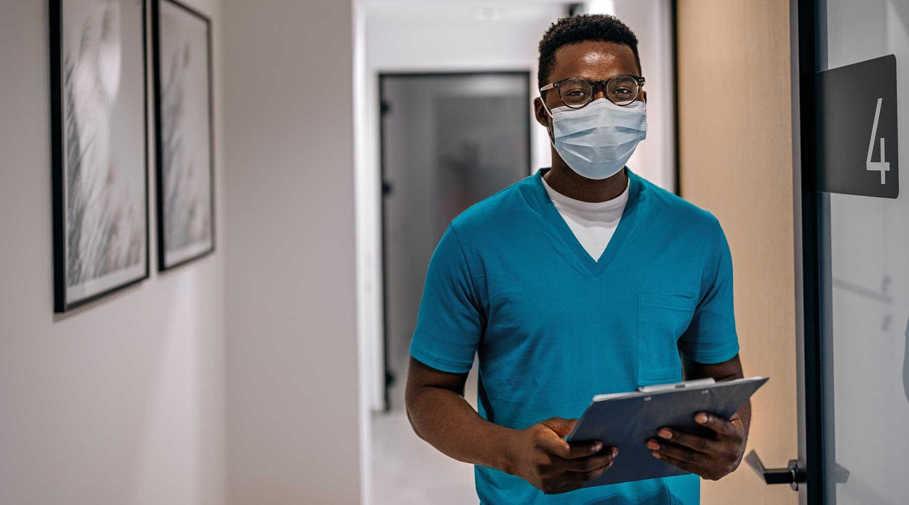 Male nurse practitioner in blue scrubs and face mask holding a clipboard in hospital hallway