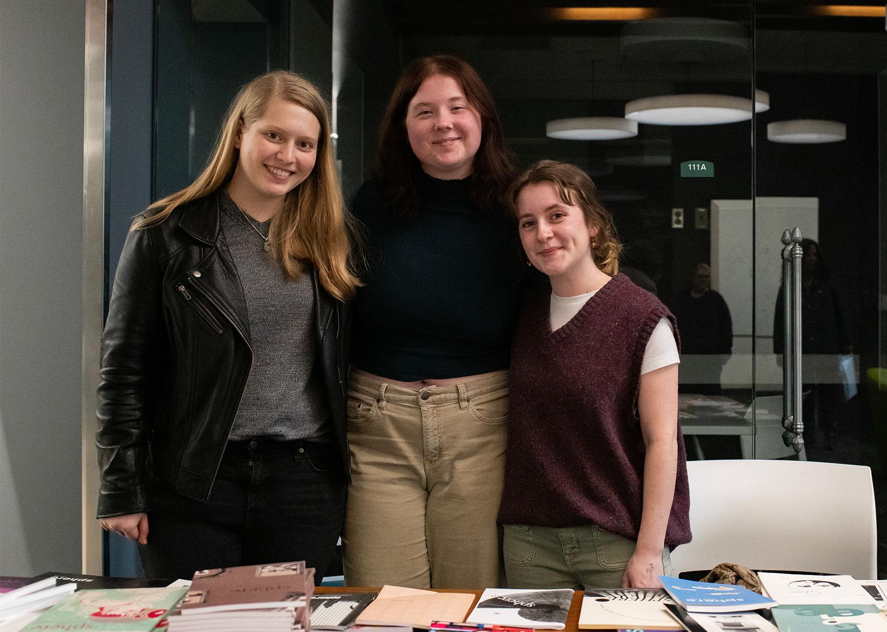 Three students pose for a photo in behind a table with books on it in Alden Library.
