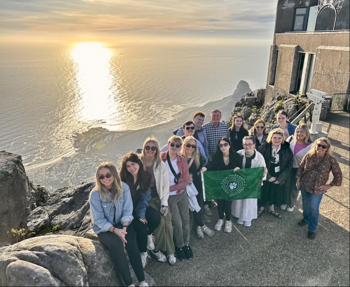 A group of students and faculty pose with an OHIO flag with the South Africa coast line in the background. 